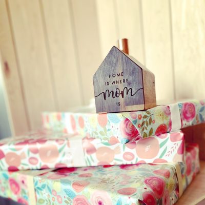Une petite maison en bois avec une cheminée en cuivre est posée sur une pile de cadeaux sur lesquels sont gravés les mots "La maison, c'est là où se trouve maman".