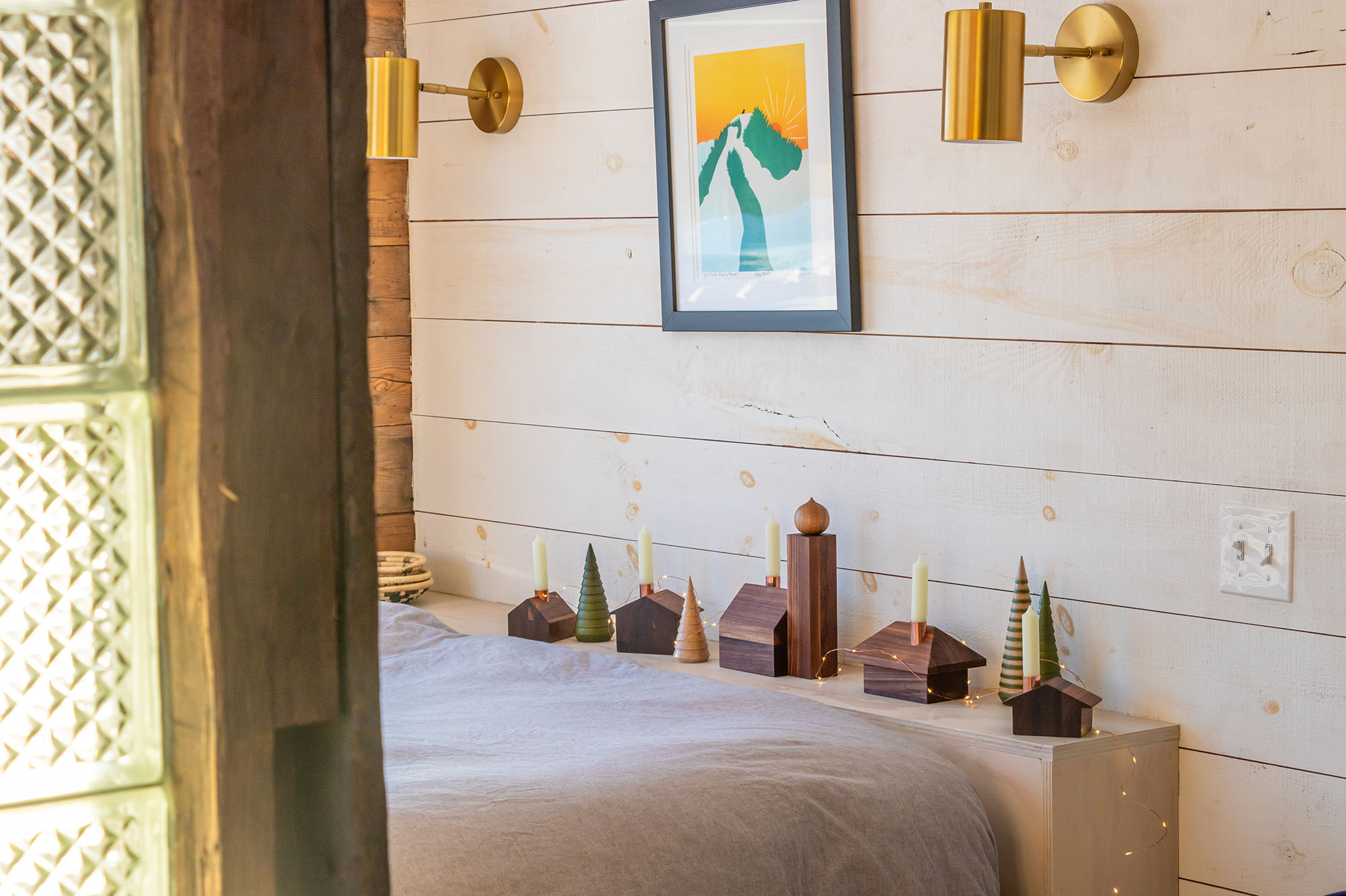 A wooden alpine village display sits on a light wooden surface against a shiplap wall. Copper chimneys hold unlit candles, while fairy lights weave through the arrangement, creating a cozy, festive ambiance.