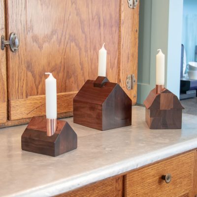 Un ensemble de trois minuscules chandeliers en bois posés sur un comptoir, avec des bougies blanches effilées sortant de leurs cheminées en cuivre.