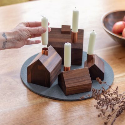 Quatre petits chandeliers en bois avec des bougies blanches effilées dans leurs cheminées en cuivre sont posés sur un tapis de feutre gris à côté d'un bol de fruits et de brindilles décoratives.