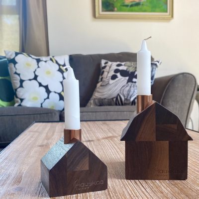 Two tiny wooden candlestick holders. One is a barn and one is a little cottage. They both have white tapered candles and they are in a living room.