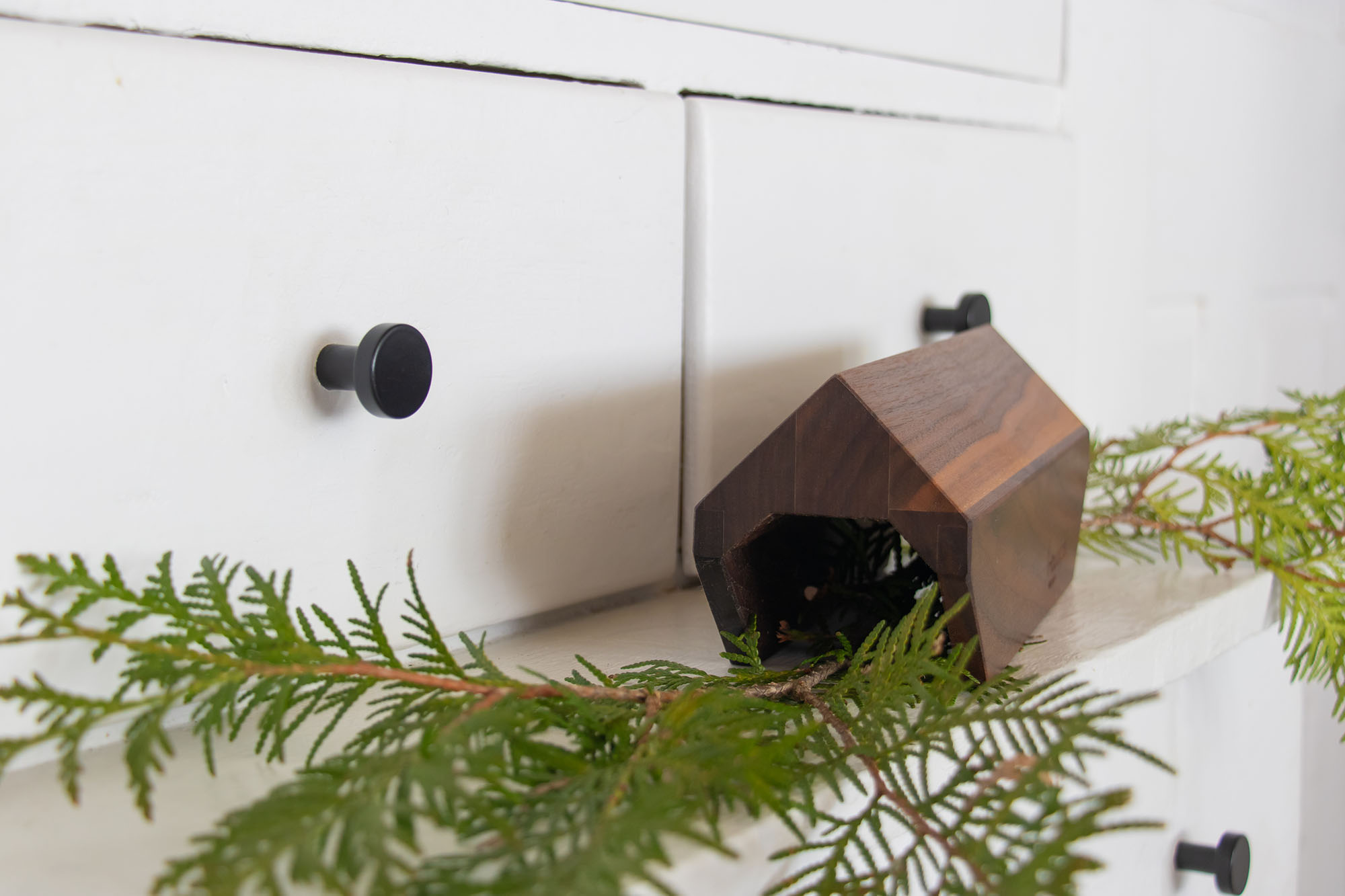 A small wooden covered bridge decor piece displayed on a white shelf, styled with evergreen branches in front of white cabinetry with black knobs.