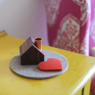 Une petite maison en bois avec une cheminée en cuivre est posée sur un bloc de feutre gris circulaire avec un cœur en feutre rouge placé à côté.