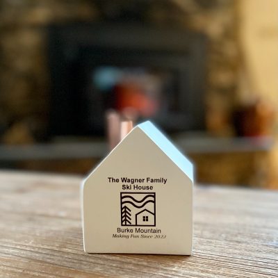 Un petit bougeoir en bois blanc avec une cheminée en cuivre, une image décorative gravée sur mesure et les mots "The Wagner Family Ski House. Burke Mountain. Making Fun Since 2022" (S'amuser depuis 2022).